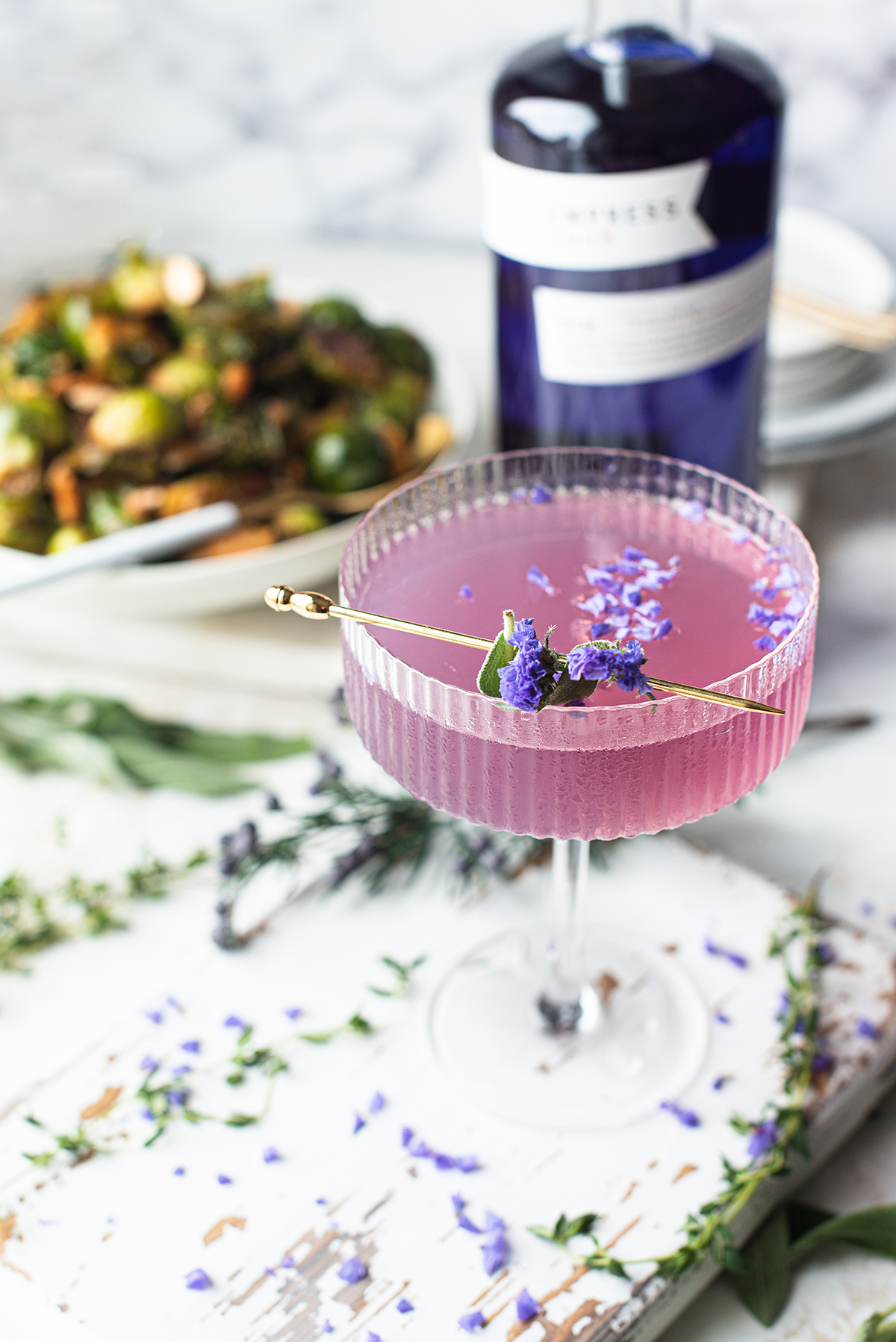 purple gin cocktail sprinkled with purple flowers on white board with Brussels sprouts & Empress Gin