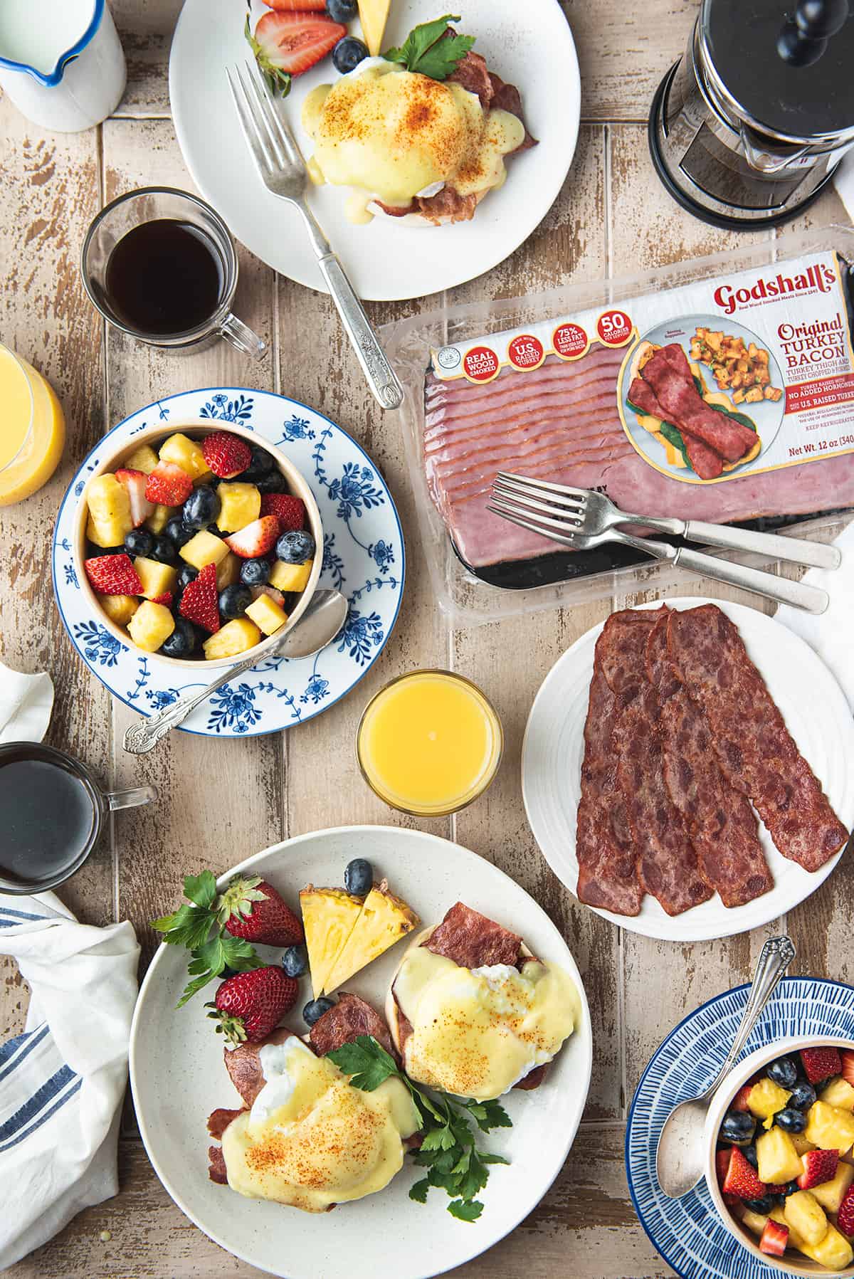 breakfast spread on wooden table with plates of eggs Benedict, bacon, bowls of fruit, and orange juice