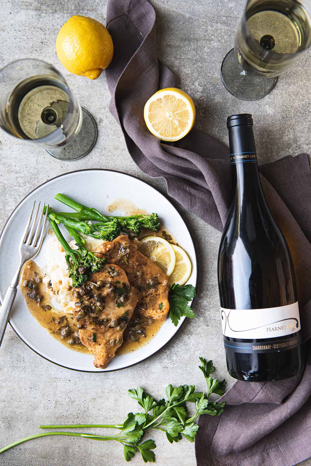 white plate with two chicken piccata breasts, mashed potatoes, greens + bottle of white wine