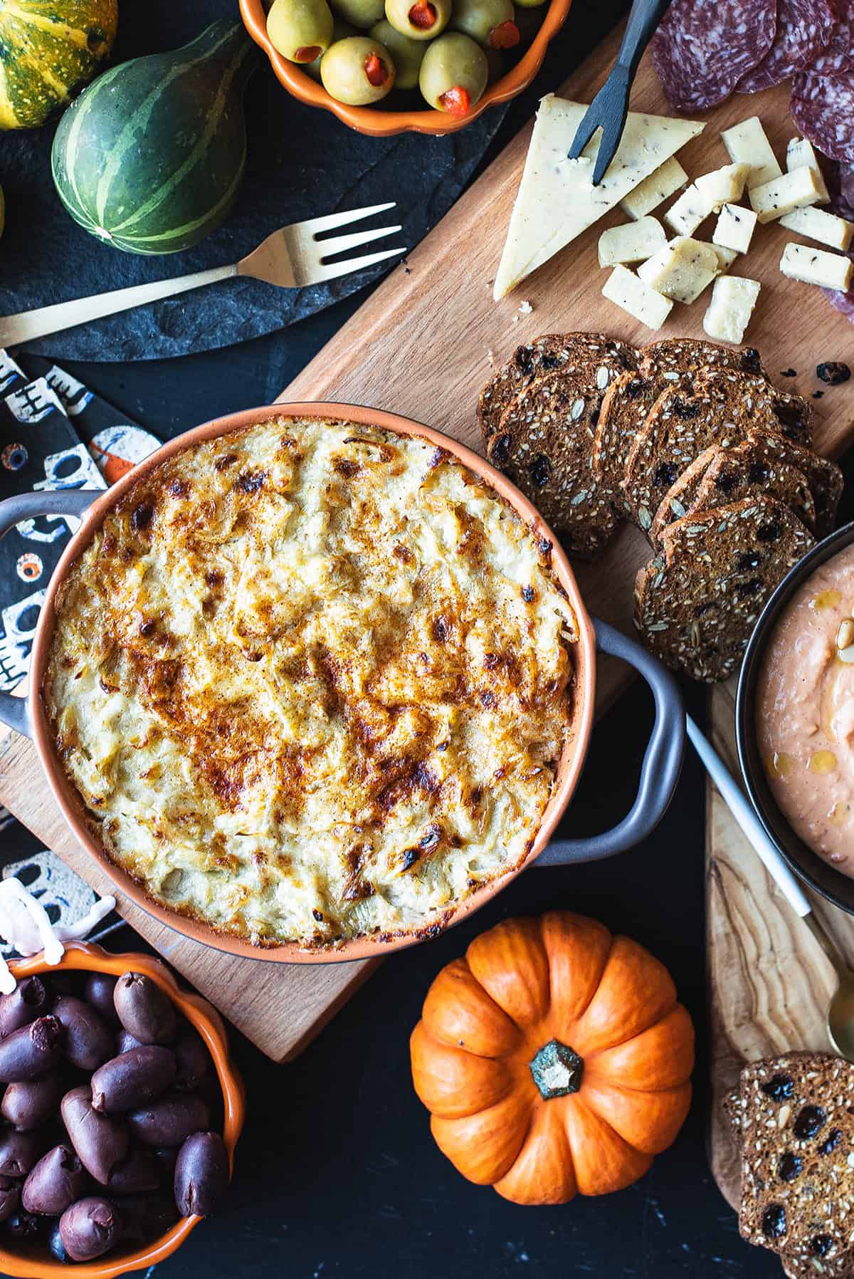Easy artichoke dip recipe served in a baking dish on a wooden board with bread and olives