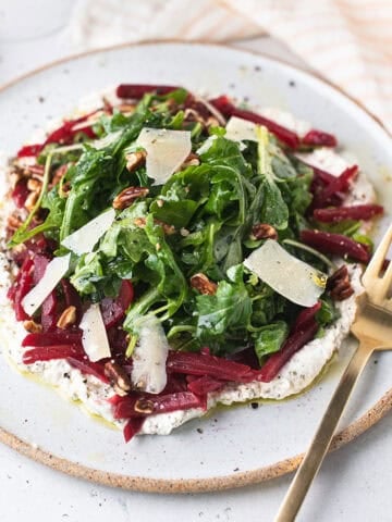 arugula beet salad with herbed ricotta, julienned beets and shaved parmesan on a speckled white plate with gold fork