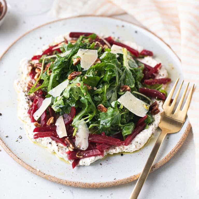 arugula beet salad with herbed ricotta, julienned beets and shaved parmesan on a speckled white plate with gold fork