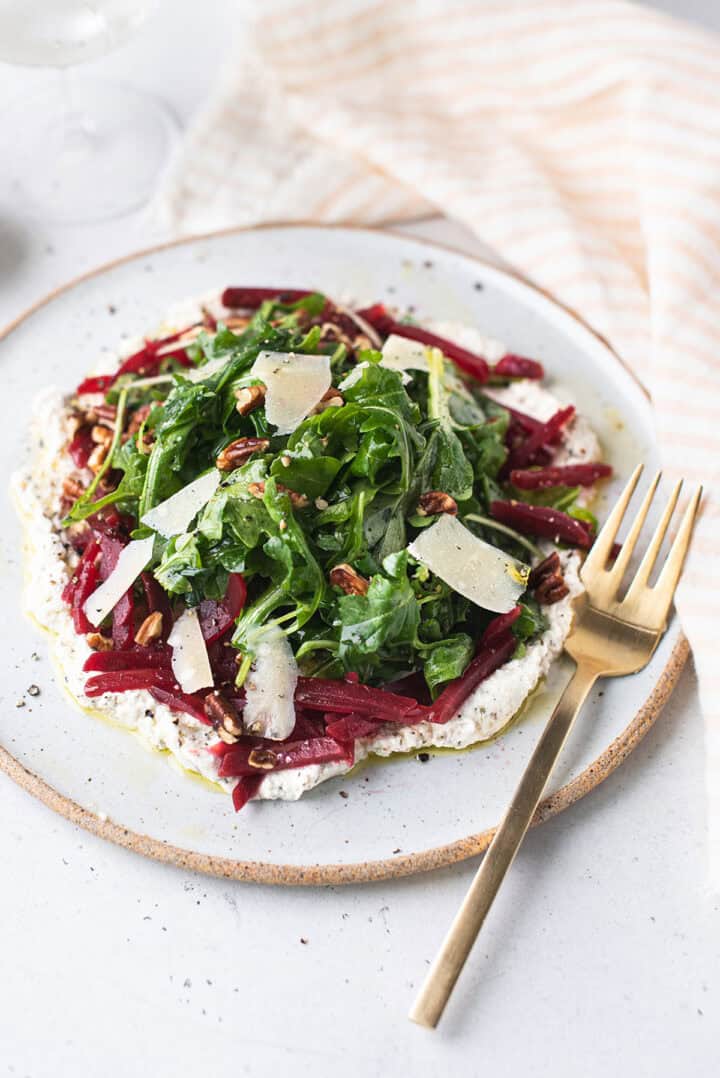 arugula beet salad with herbed ricotta, julienned beets and shaved parmesan on a speckled white plate with gold fork