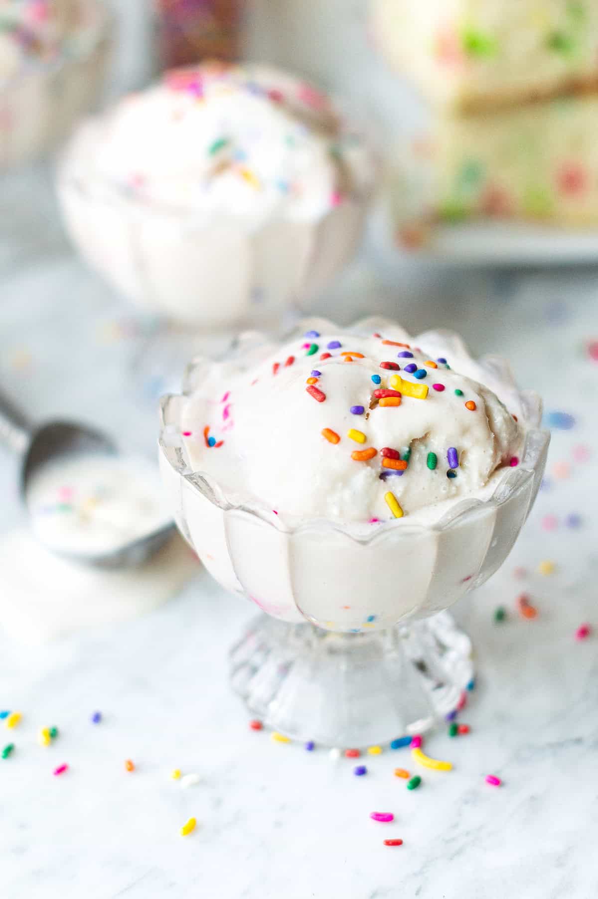 close-up of birthday cake ice cream in a glass dish topped with sprinkles, birthday cake in background