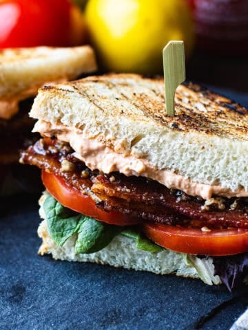 BLT sandwich halves on a slate platter with grilled sourdough, stacked candied bacon, lettuce, and tomato with a Sriracha bottle in the background