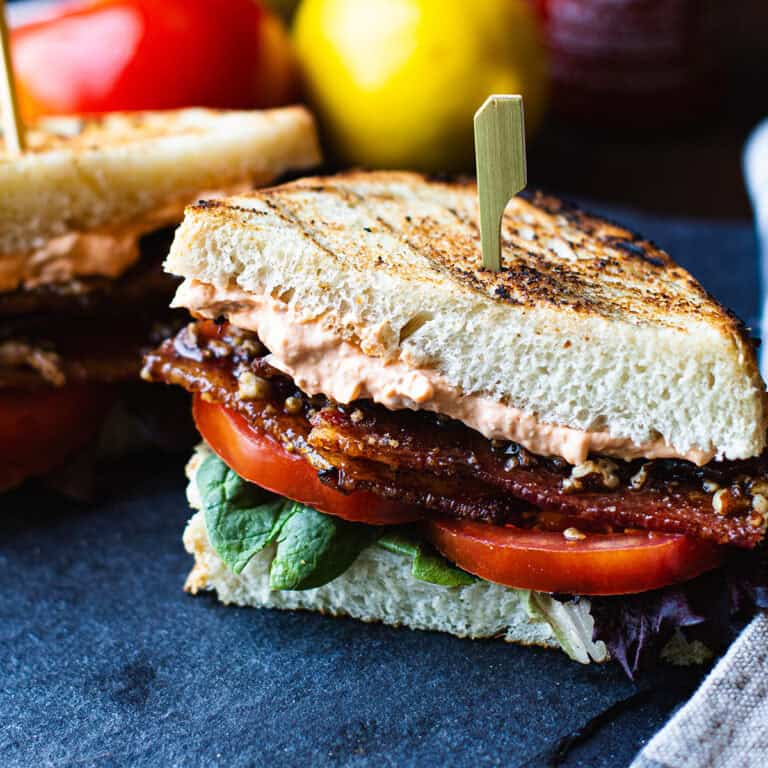 BLT sandwich halves on a slate platter with grilled sourdough, stacked candied bacon, lettuce, and tomato with a Sriracha bottle in the background