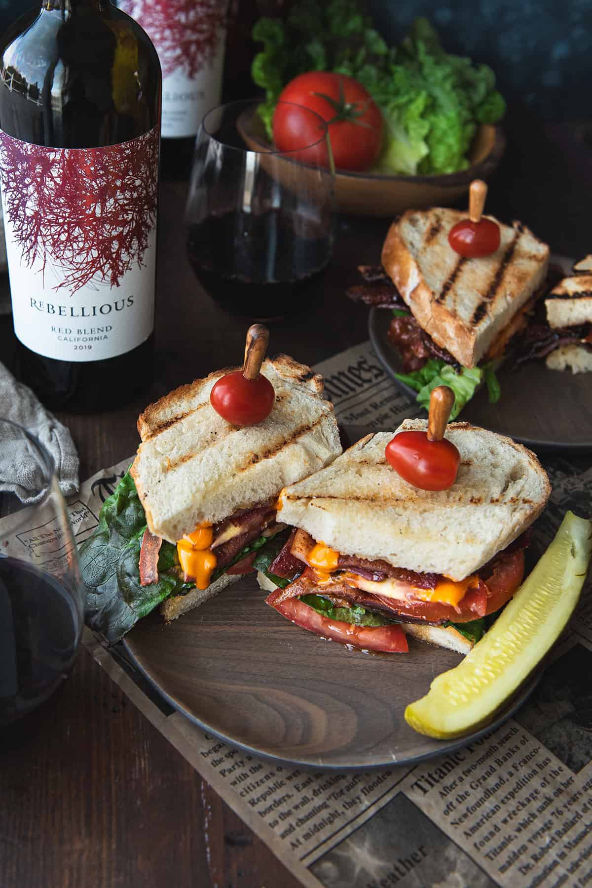 plated BLT sandwiches with pickle spears, cherry tomato sandwich picks, a glass of red wine, and a bowl of lettuce and tomato on a wooden table