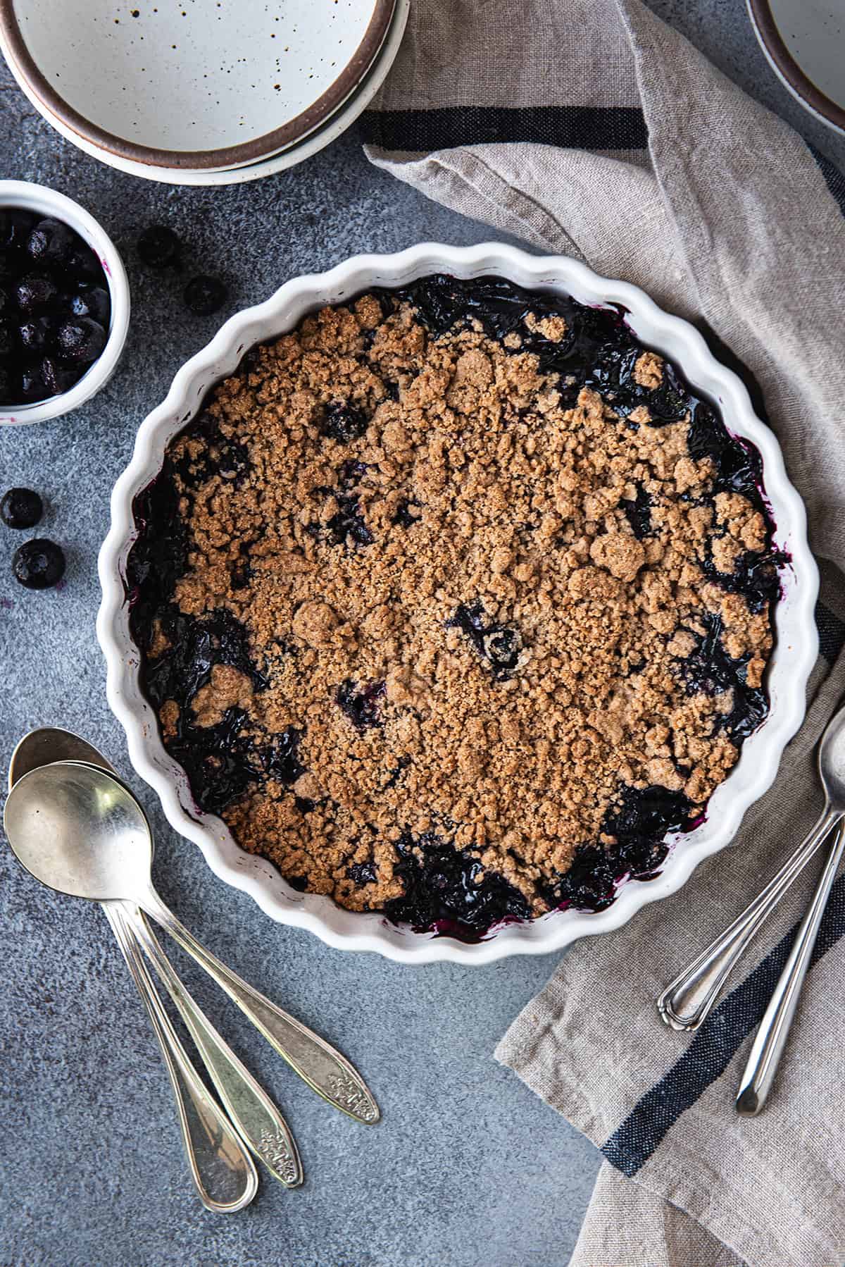 Blueberry crumble with a golden brown streusel topping in a white fluted baking dish, with a small bowl of fresh blueberries and vintage spoons beside it.