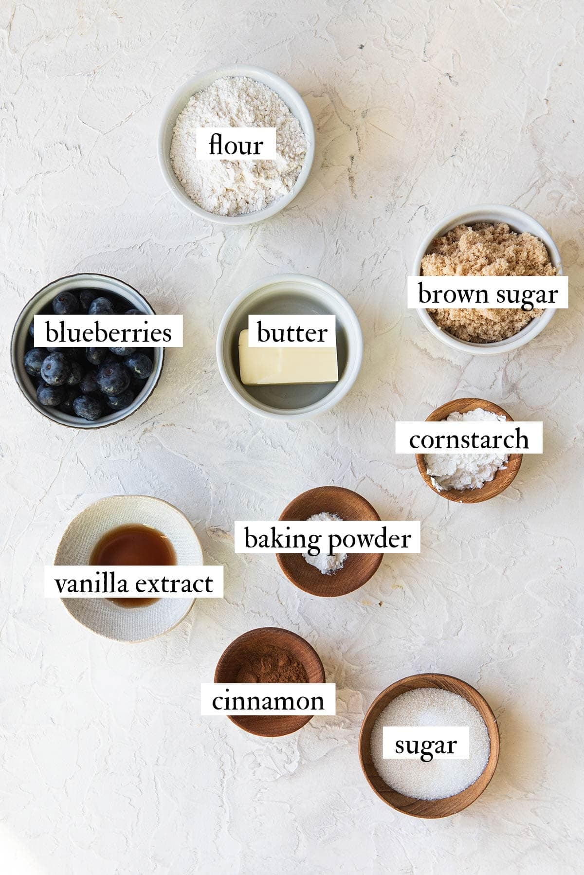 Labeled ingredients for blueberry crumble in small bowls on a white surface: fresh blueberries, flour, butter, brown sugar, sugar, cornstarch, baking powder, cinnamon, and vanilla extract.