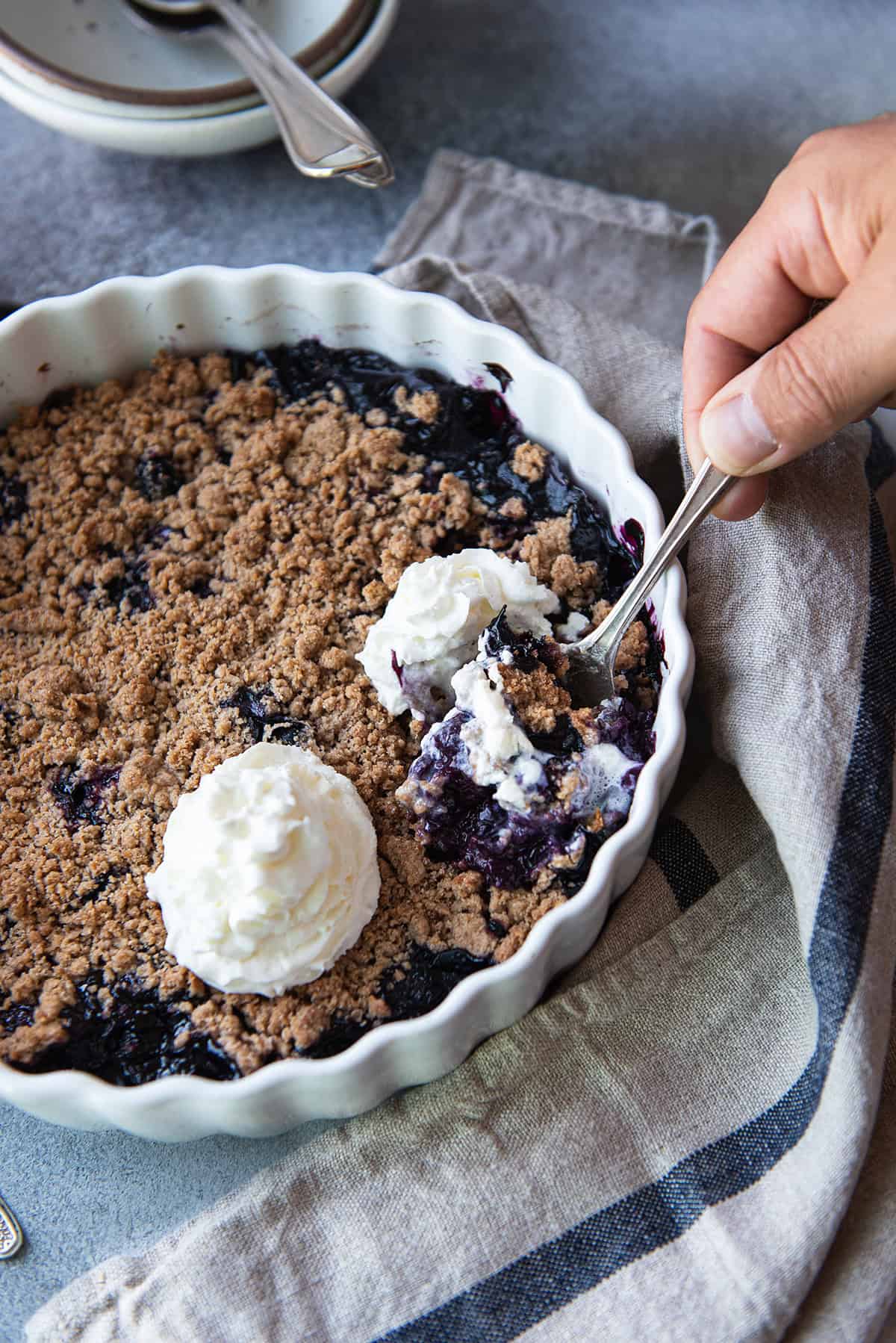 A hand spooning a serving of blueberry crumble with whipped cream, showing the thick blueberry filling under the crisp streusel topping.