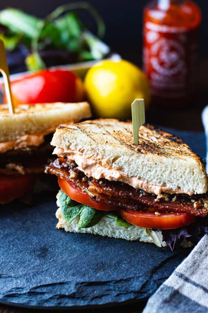 Two BLT sandwich halves on a slate platter with grilled sourdough, caramelized candied bacon, fresh tomato, spring mix, and a visible layer of Sriracha aioli, with a Sriracha bottle and lemon blurred in the background