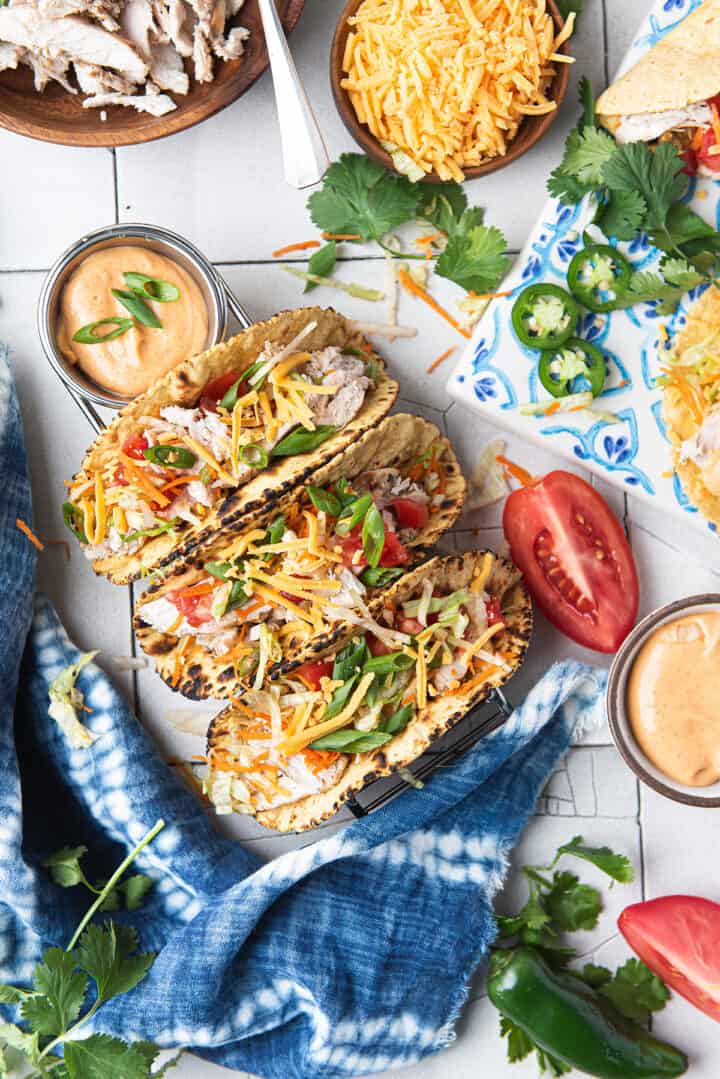three chicken tacos, topped with lettuce, tomatoes, and cheese on white table with blue napkin