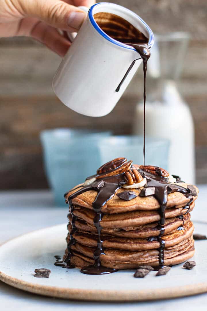 hand pouring chocolate syrup over stack of chocolate pancakes on white plate
