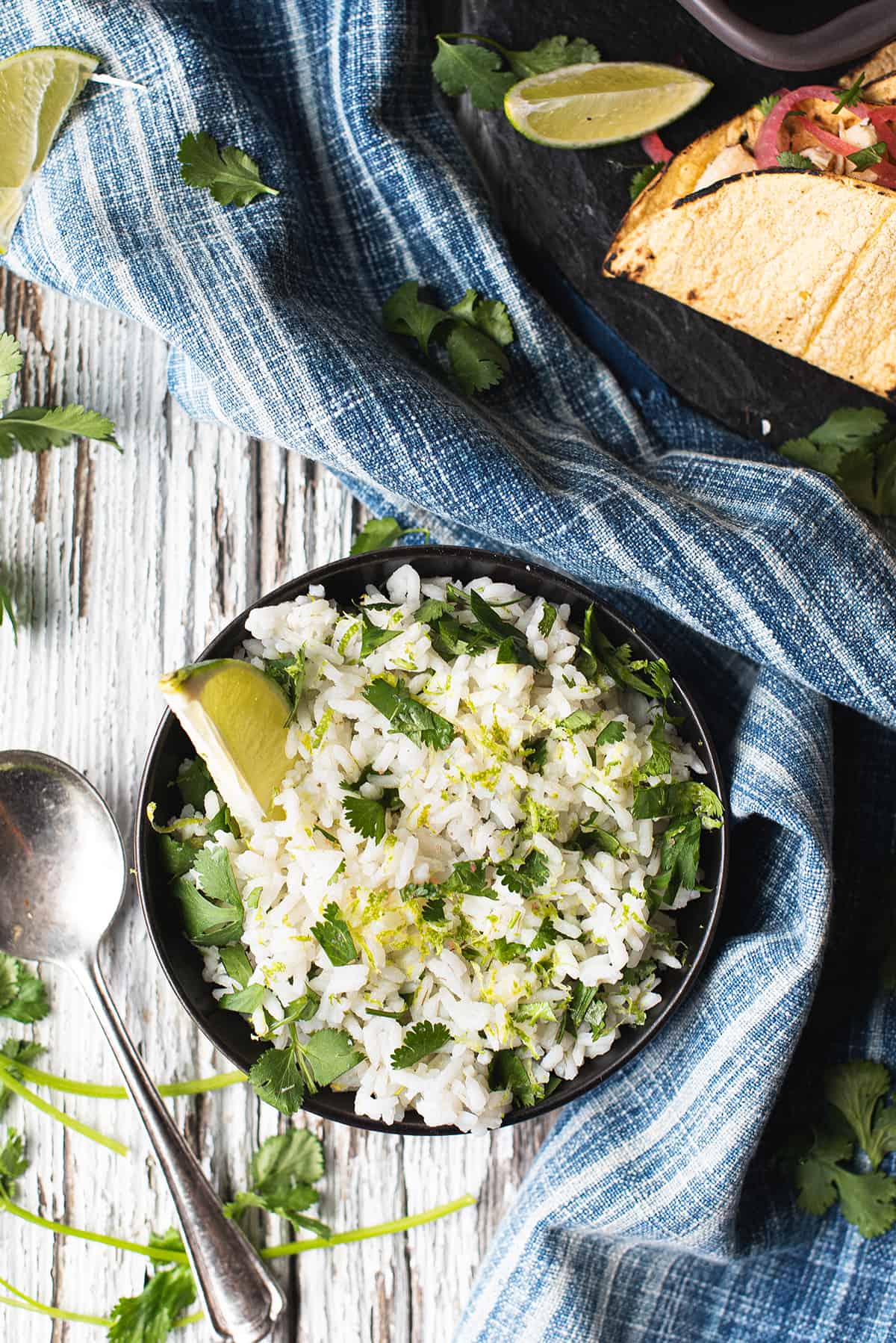cilantro lime rice in black bowl with lime wedge on white wooden table with spoon + blue napkin