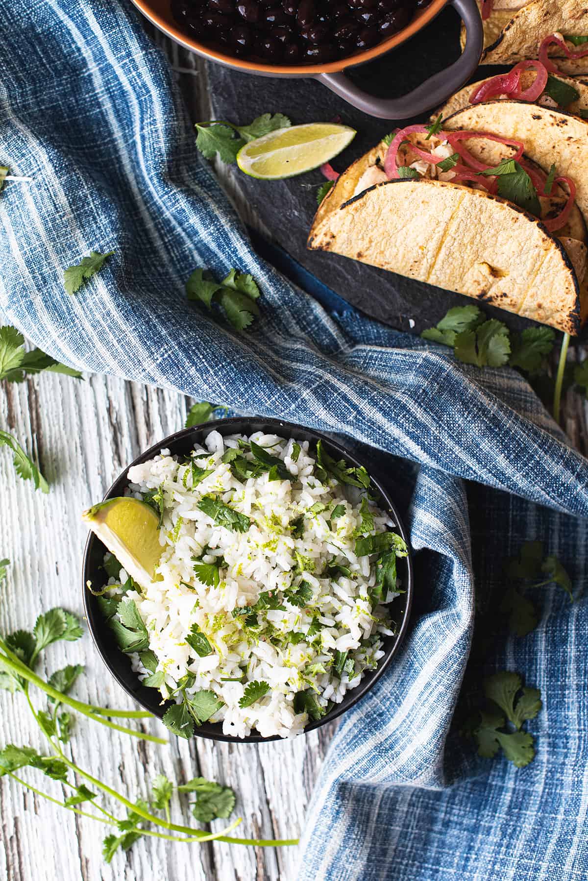 cilantro lime rice in black bowl on white wooden table with plate of tacos, lime wedges, blue napkin
