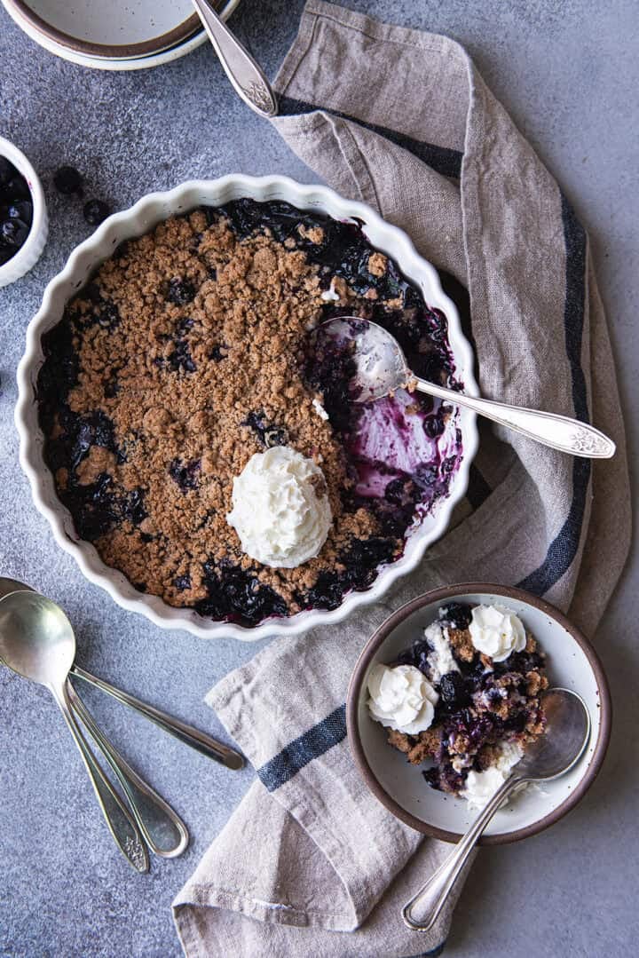 Overhead view of blueberry crumble with a portion scooped out to show the jammy berry filling, topped with whipped cream, next to a serving in a stoneware bowl.
