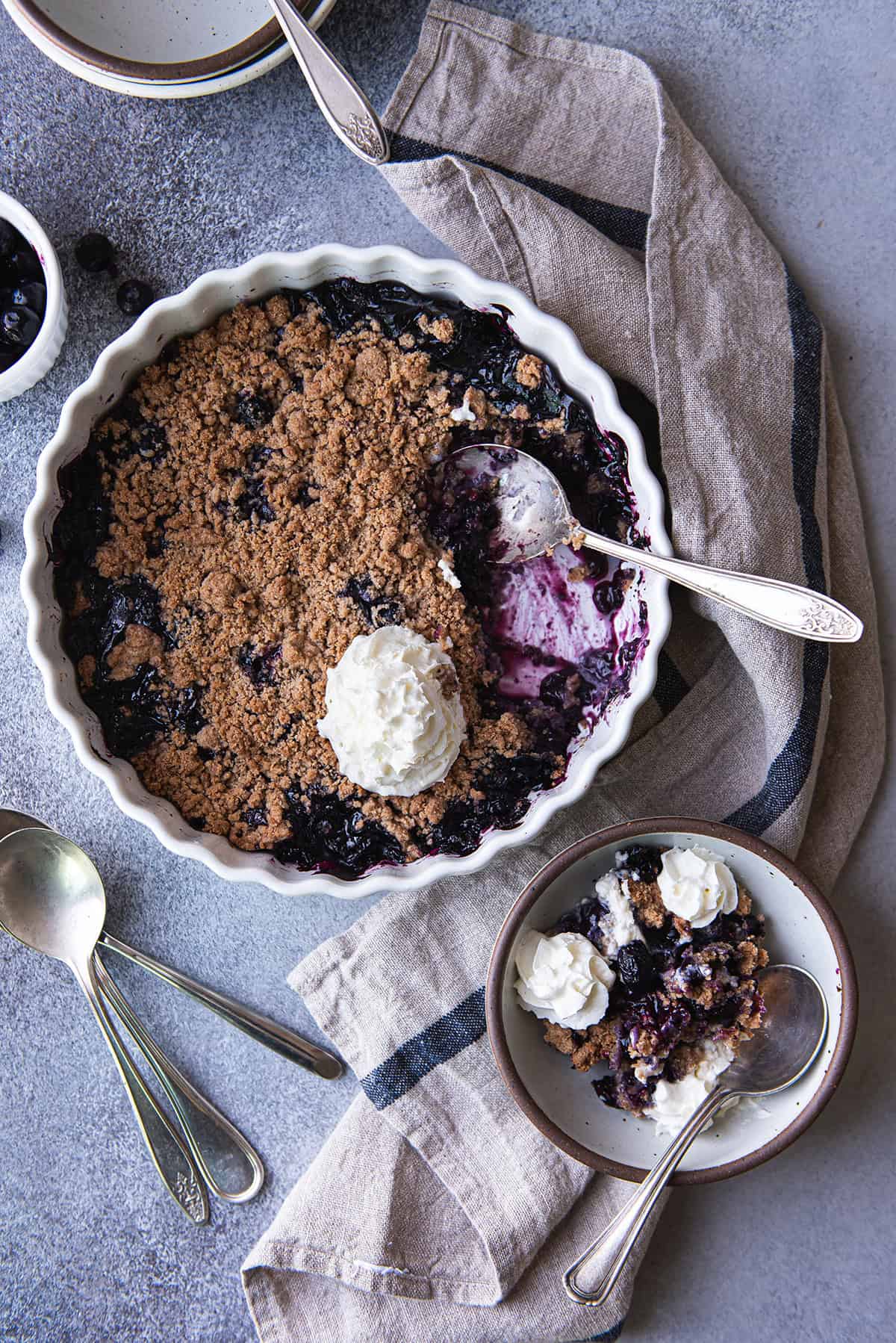Overhead view of blueberry crumble with a portion scooped out to show the jammy berry filling, topped with whipped cream, next to a serving in a stoneware bowl.