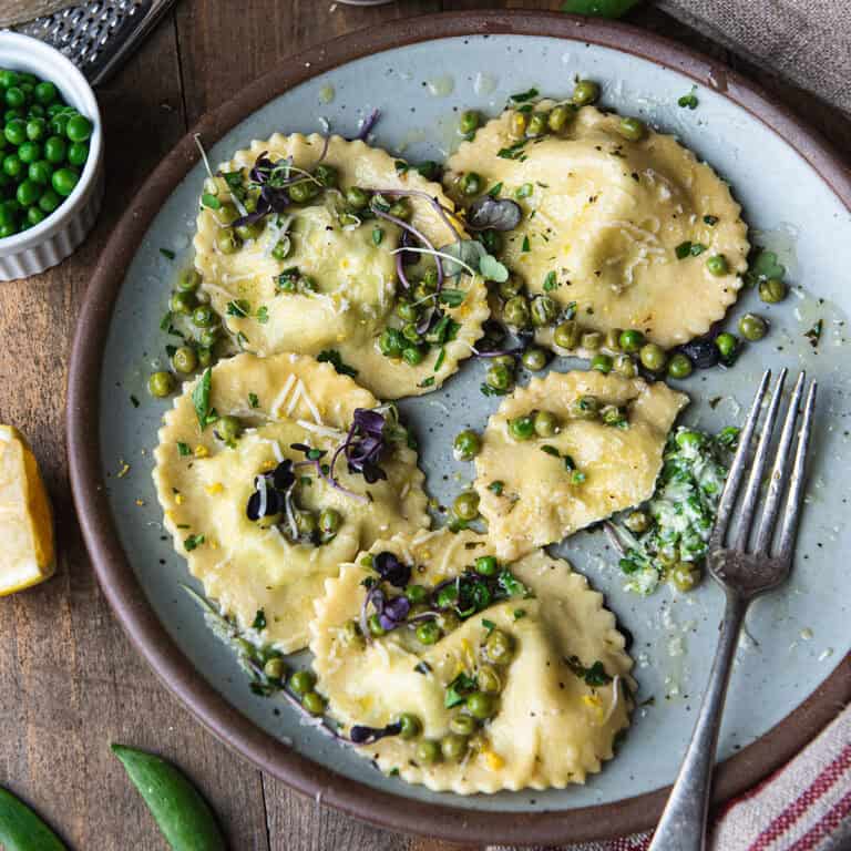 homemade ricotta ravioli with peas in lemon butter sauce on a blue ceramic plate, garnished with microgreens and shaved parmesan
