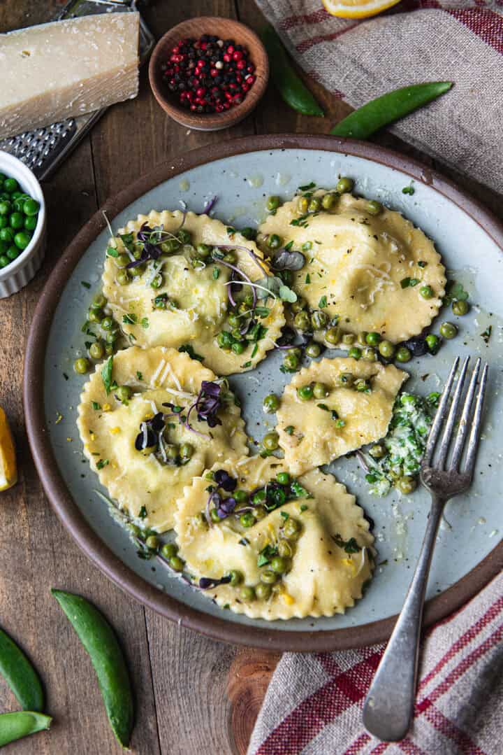 homemade ricotta ravioli with peas in lemon butter sauce on a blue ceramic plate, garnished with microgreens and shaved parmesan, on a rustic wooden table