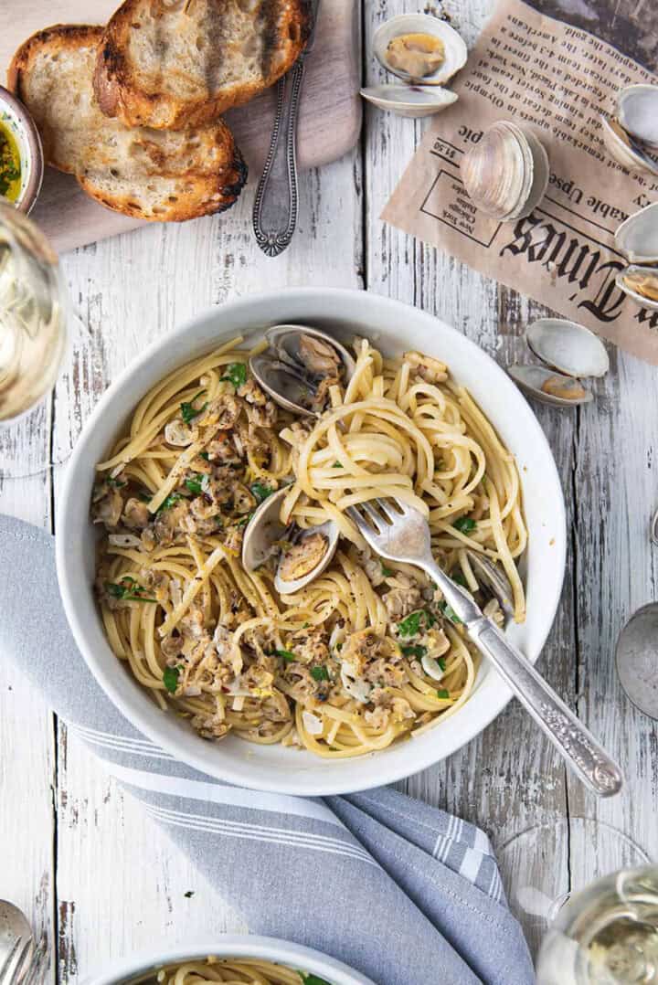 Linguine alle Vongole in a white bowl with fresh clams, chopped parsley, and a fork, served with grilled bread and white wine