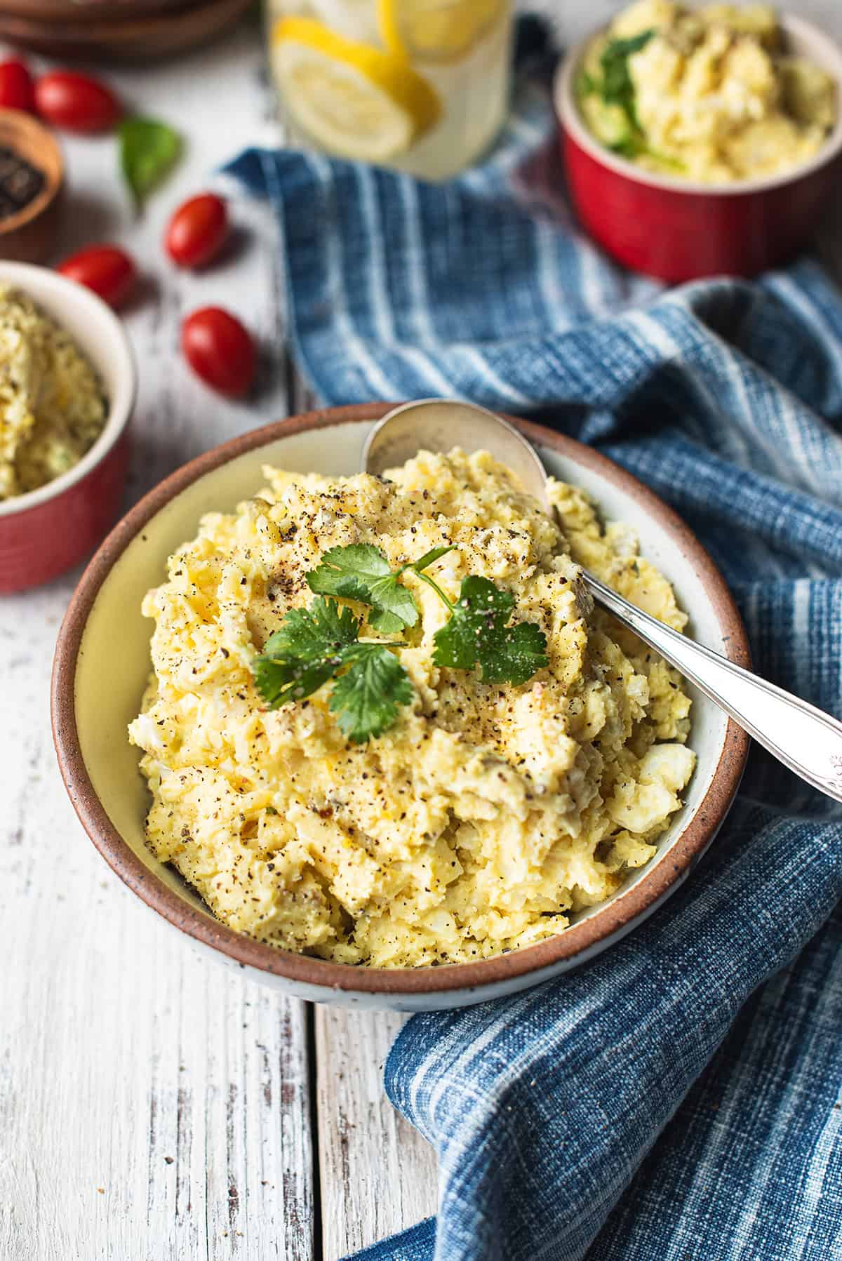 white bowl of mashed potato salad with serving spoon, garnished with parsley leaves + blue napkin