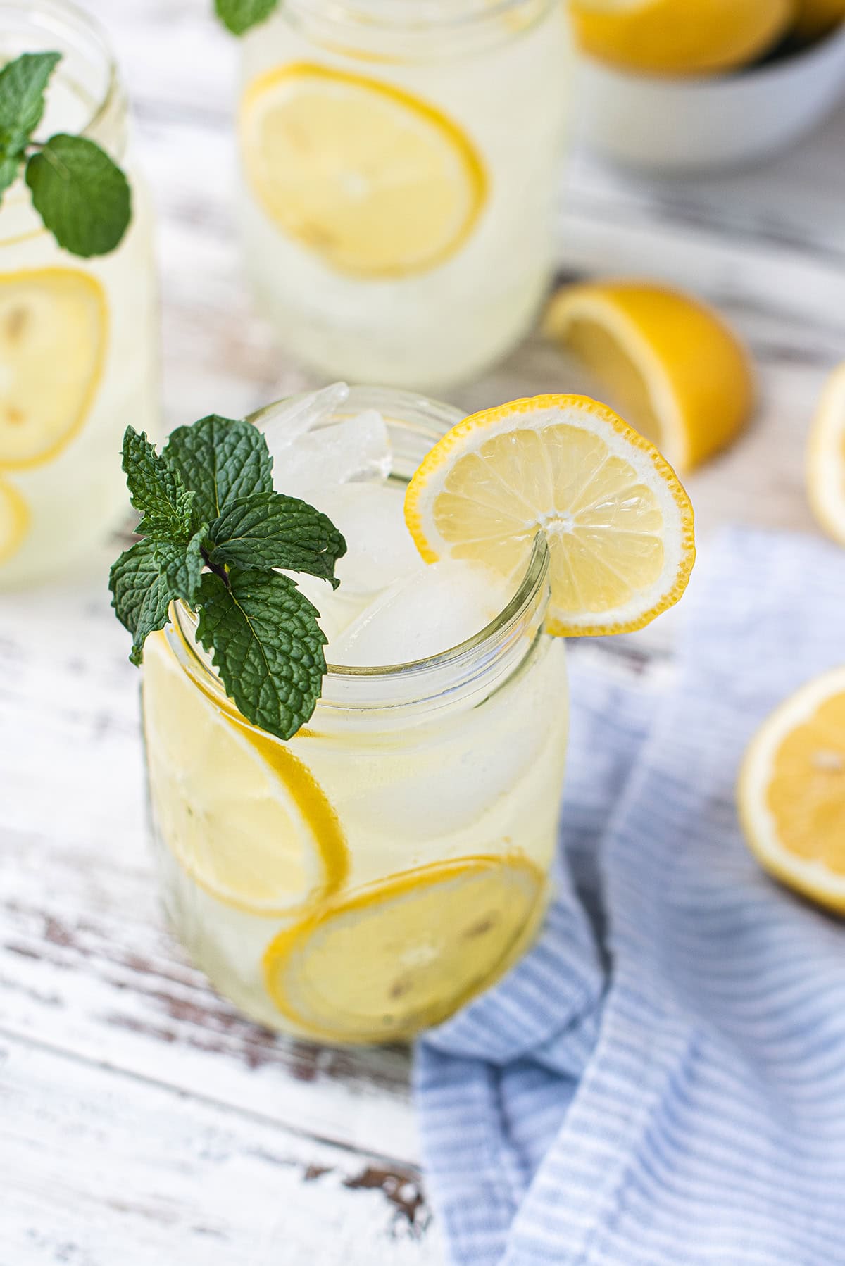 mason jars filled with lemonade, garnished with lemon slices and mint leaves, on white picnic table + blue napkin