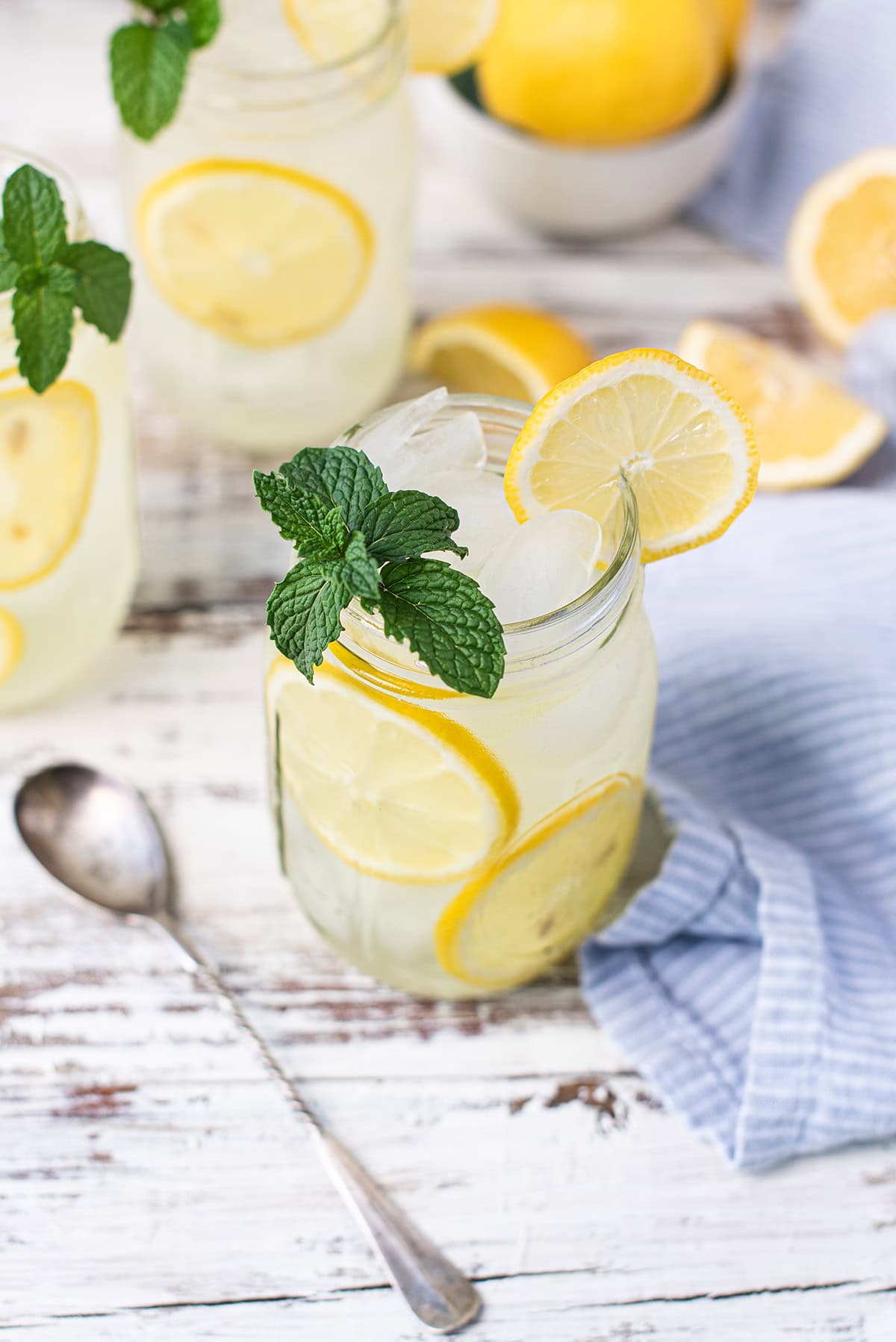 mason jars filled with mint lemonade, garnished with lemon slices and mint leaves, on white picnic table + spoon and blue napkin