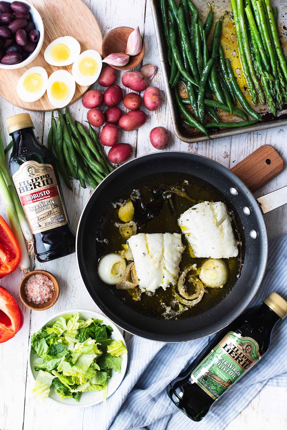 poached cod in pan surrounded by vegetables and salad ingredients on white table