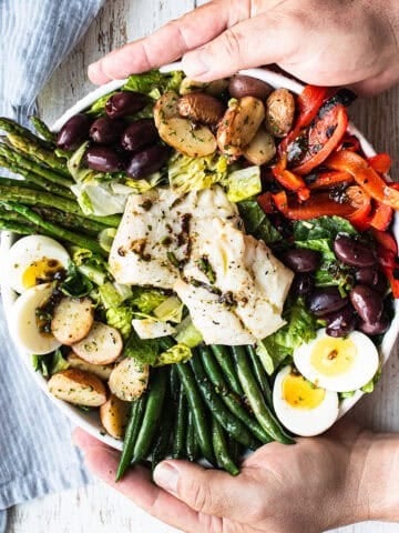 hands holding white bowl of olive oil poached cod salad, full of colorful vegetables