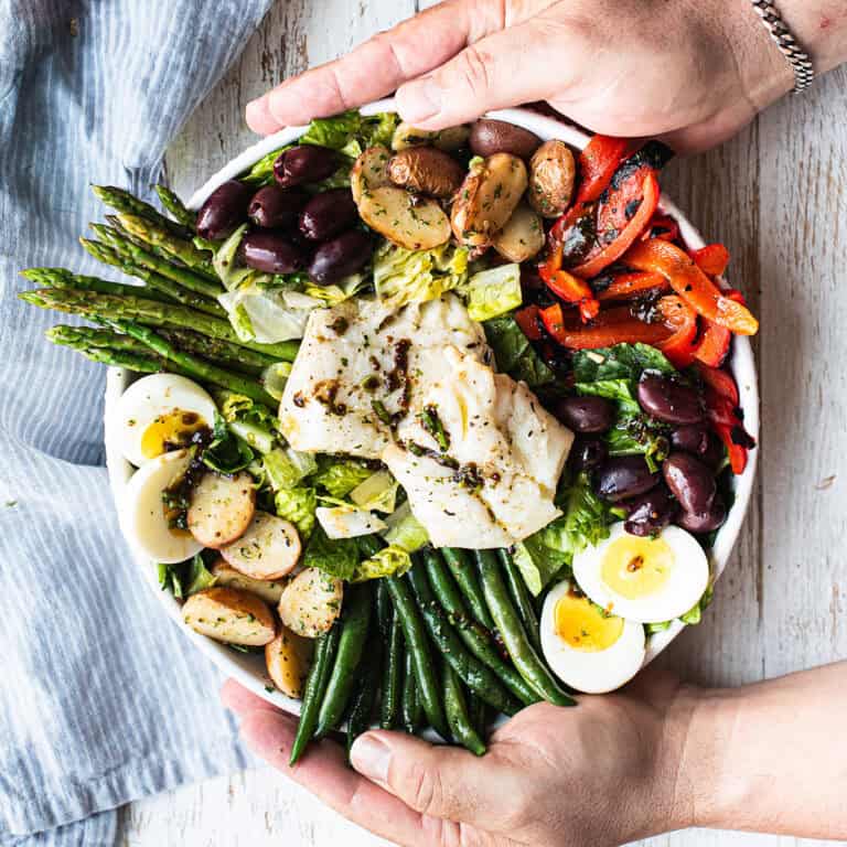 hands holding white bowl of olive oil poached cod salad, full of colorful vegetables