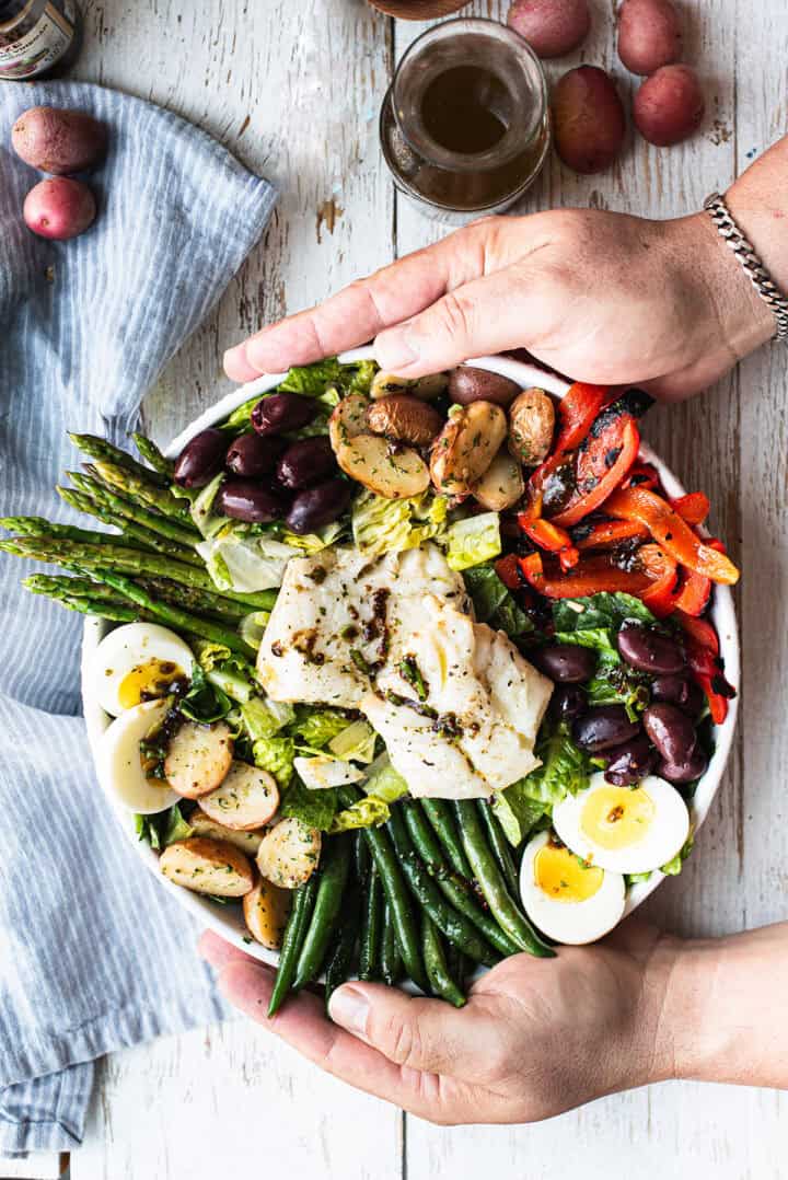 hands holding white bowl of olive oil poached cod salad, full of colorful vegetables