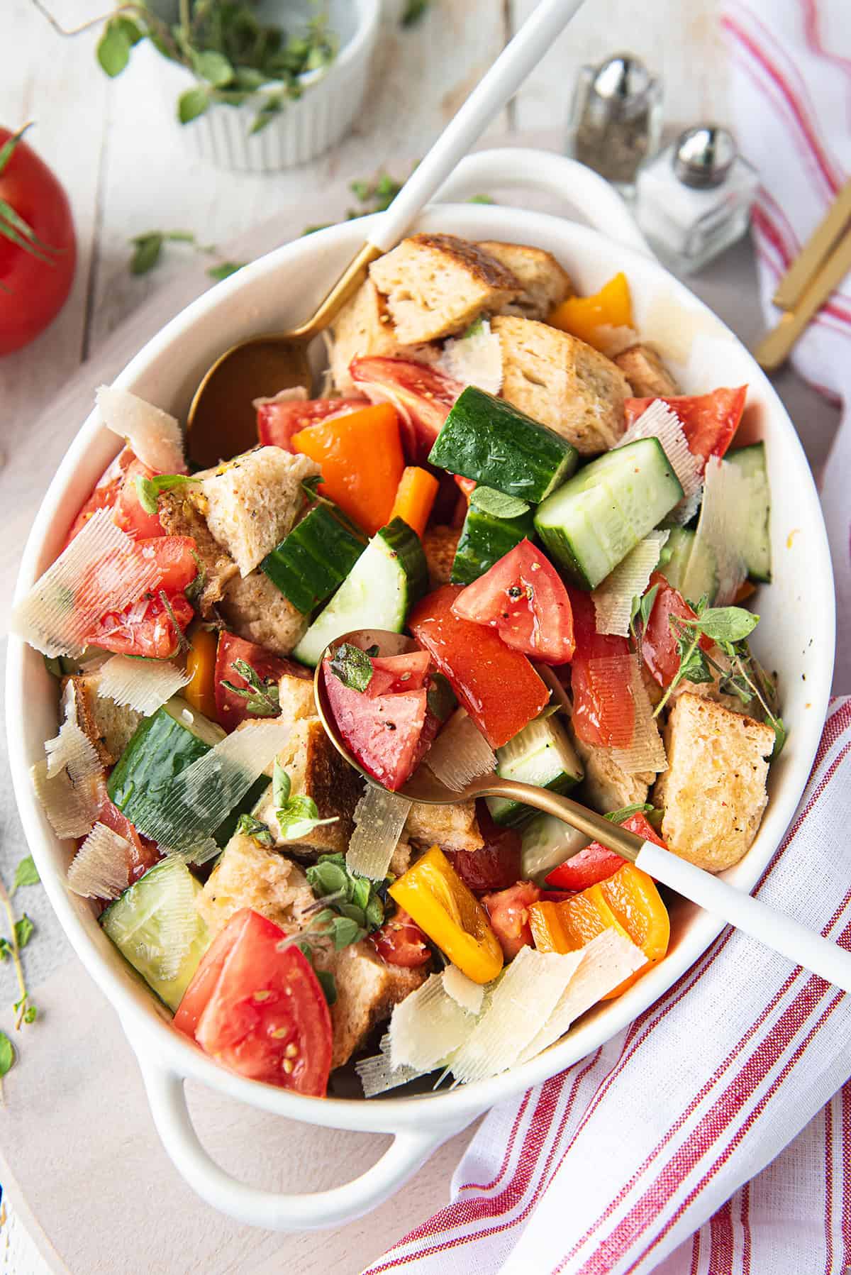 Overhead view of panzanella salad in a white oval serving bowl with tomatoes, cucumber, peppers, toasted bread, shaved Parmesan, and fresh oregano
