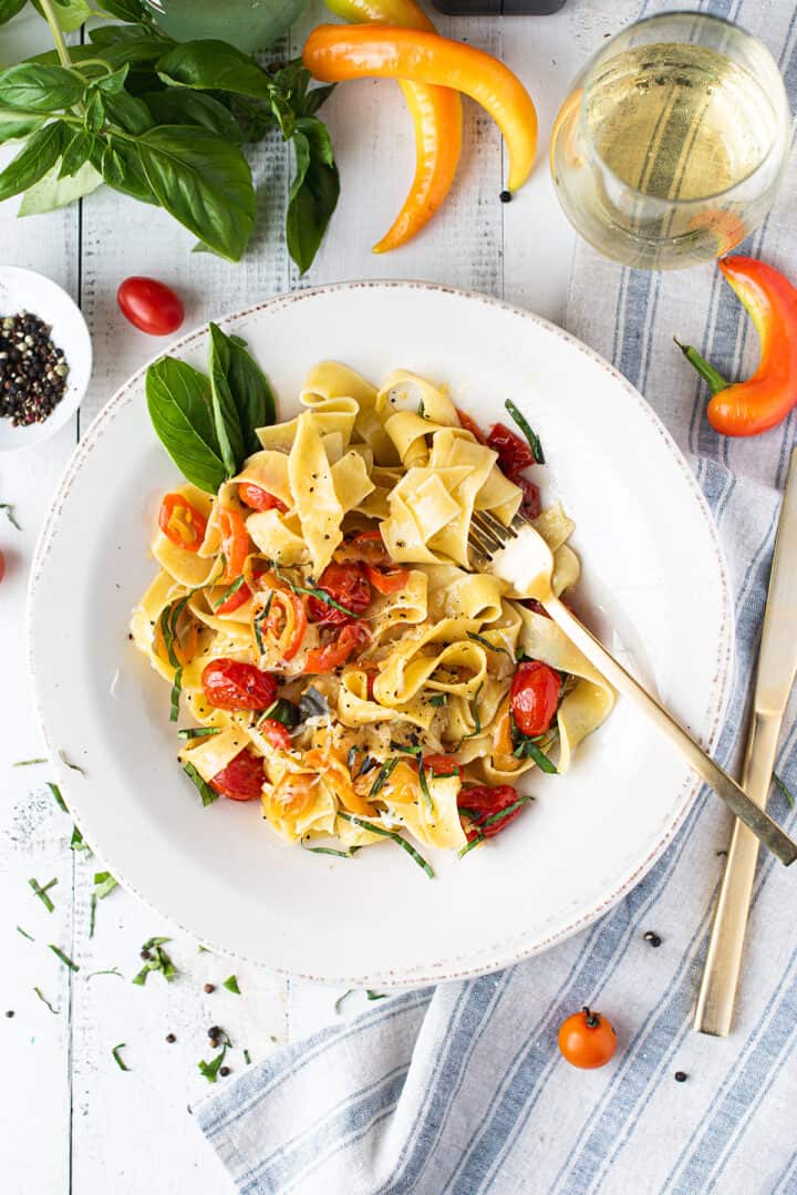 white bowl of pappardelle with tomatoes, peppers, basil on white table with blue napkin