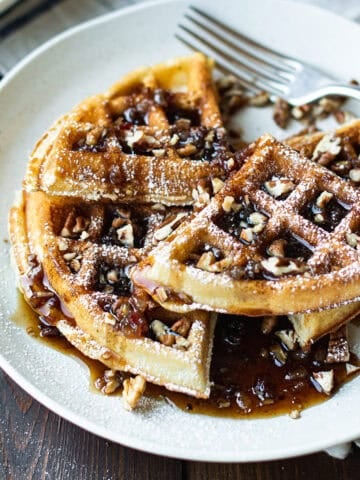 pecan waffles stacked on white plate with fork, topped with maple syrup