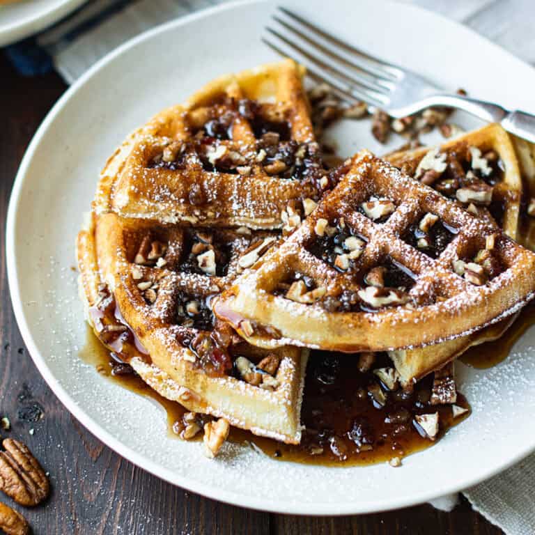 pecan waffles stacked on white plate with fork, topped with maple syrup