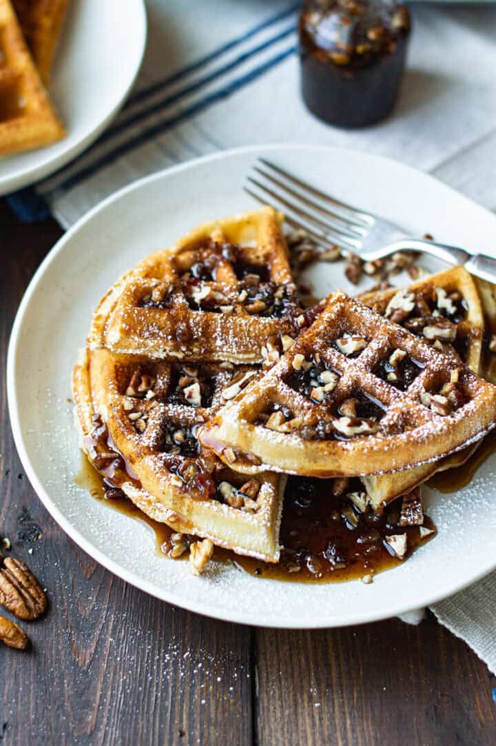 pecan waffles stacked on white plate covered in maple syrup