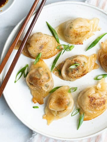 7 pork dumplings on white plate garnished with sliced green onions + chopsticks and pink striped napkin