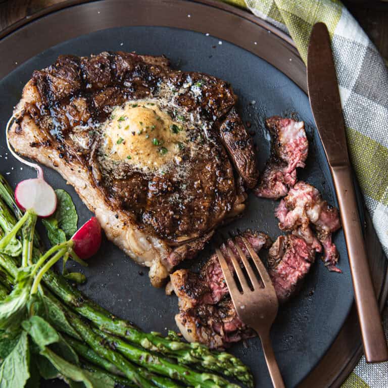 grilled ribeye steak topped with maple butter, sliced on black plate with asparagus, knife and fork