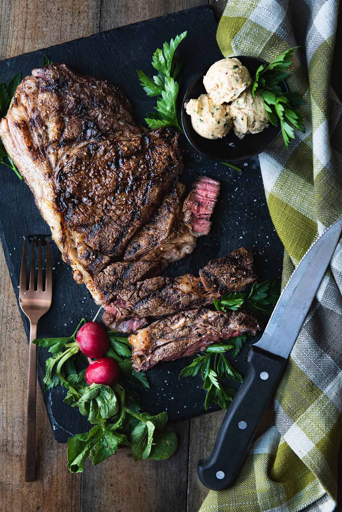 ribeye steak on black platter, sliced + steak knife, green checked napkin, and fork