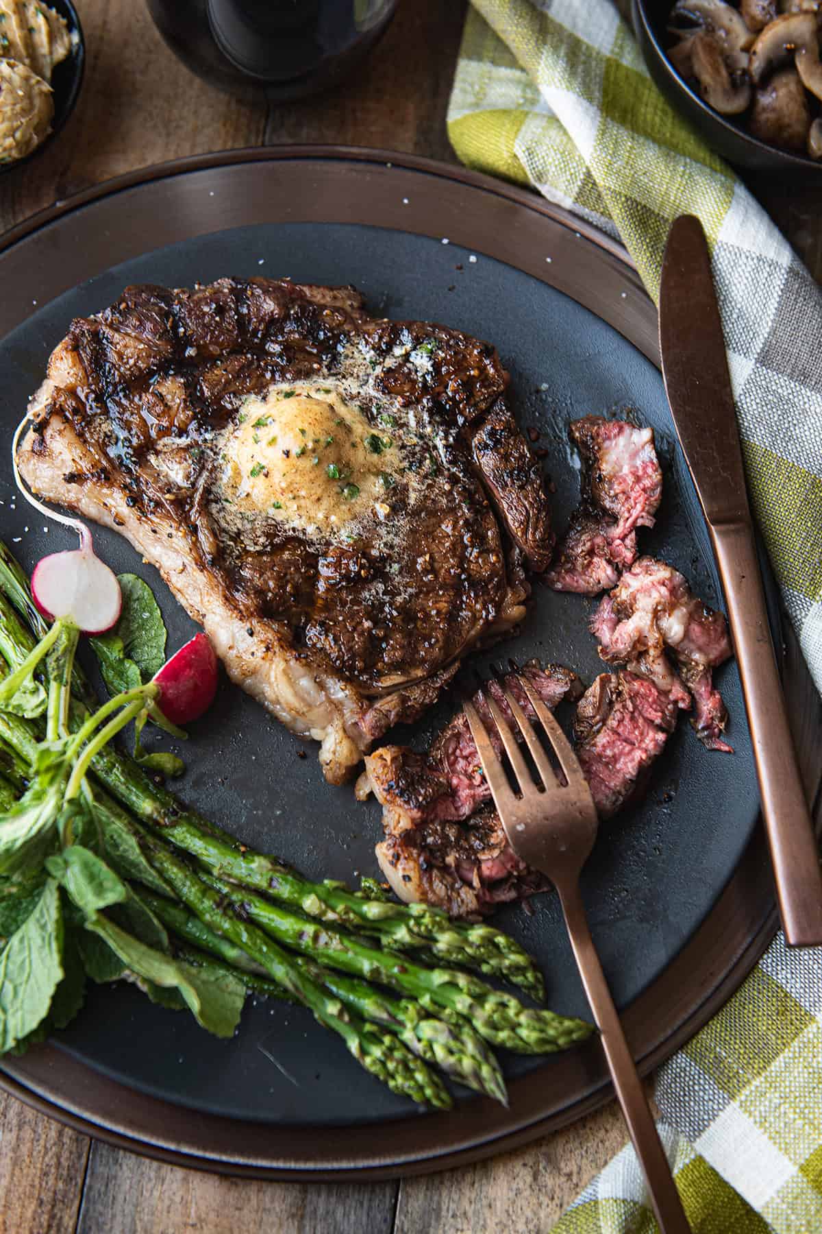 ribeye steak with melted pat of butter, sliced on black plate with asparagus, knife and fork