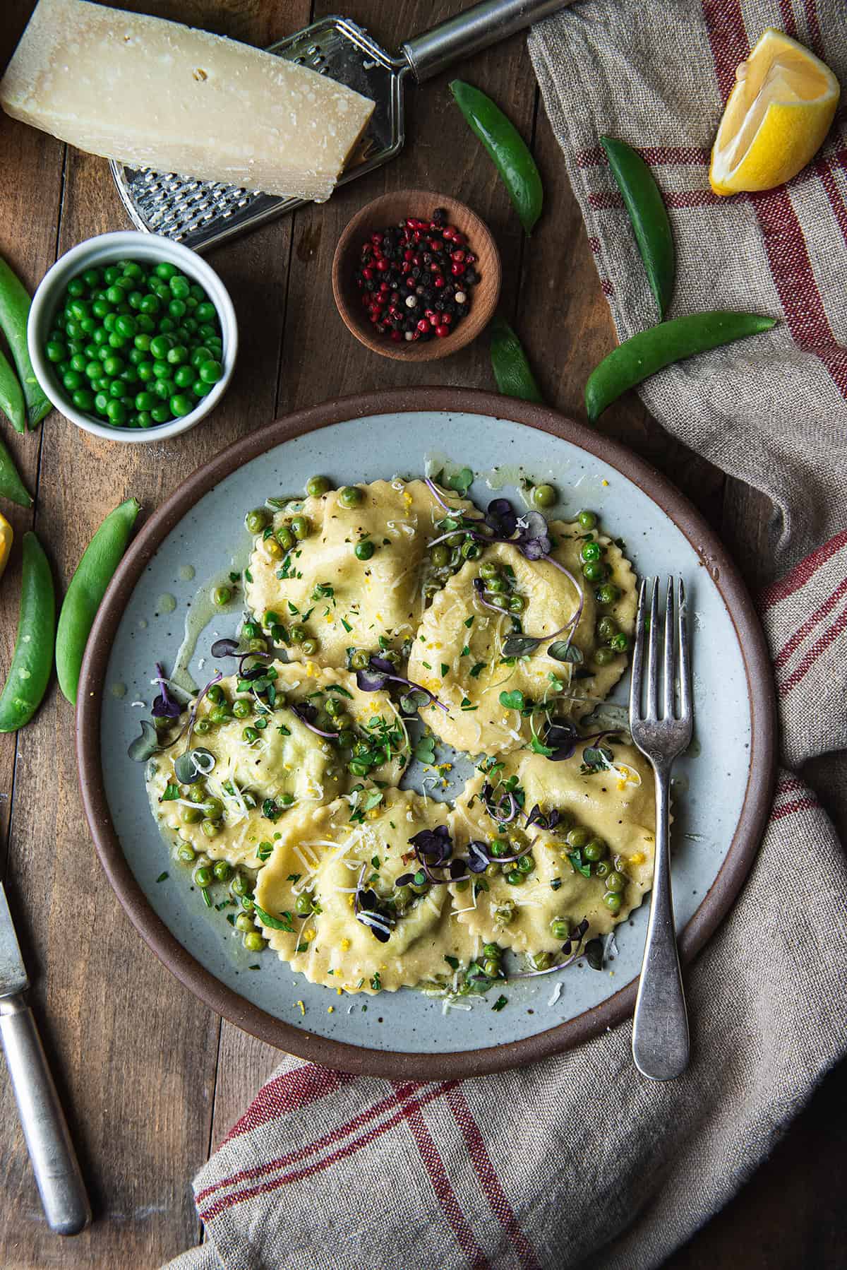 plated homemade ricotta ravioli with peas, lemon zest, and microgreens on a blue plate surrounded by fresh snap peas, parmesan, and a lemon wedge