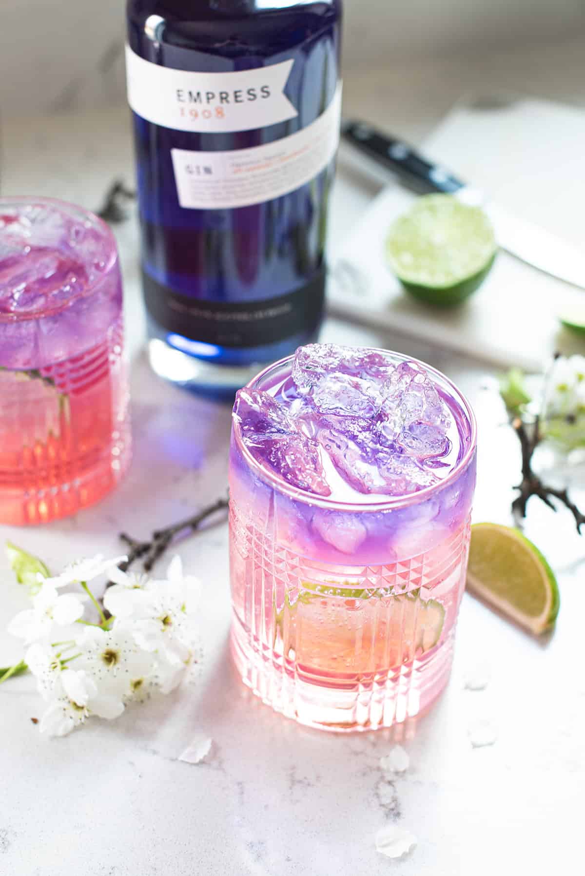 2 blue & pink empress gin cocktails on marble counter, with lime slices and blue empress bottle in background