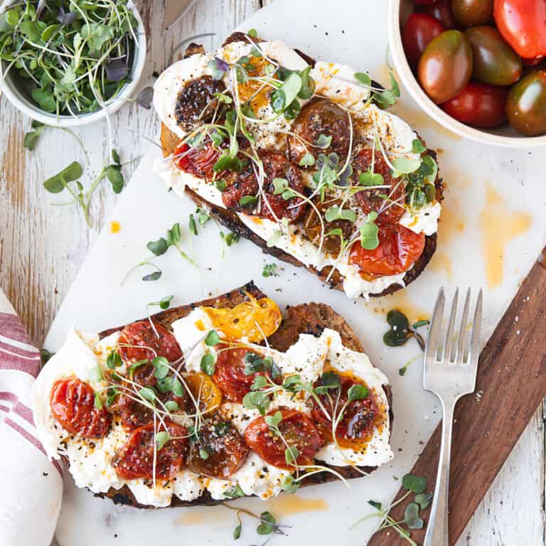 two burrata toasts on white platter, topped with roasted cherry tomatoes + fork