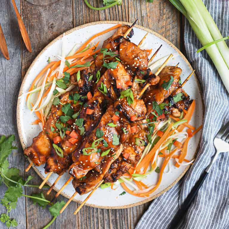 Asian BBQ chicken skewers piled on white ceramic plate with fork, blue napkin, and greens