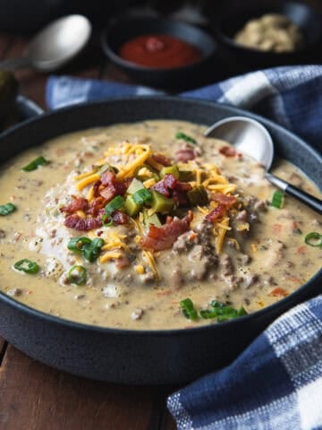 bowl of cheeseburger soup, topped chopped bacon, pickles and cheese on a rustic wooden table with blue check napkin.