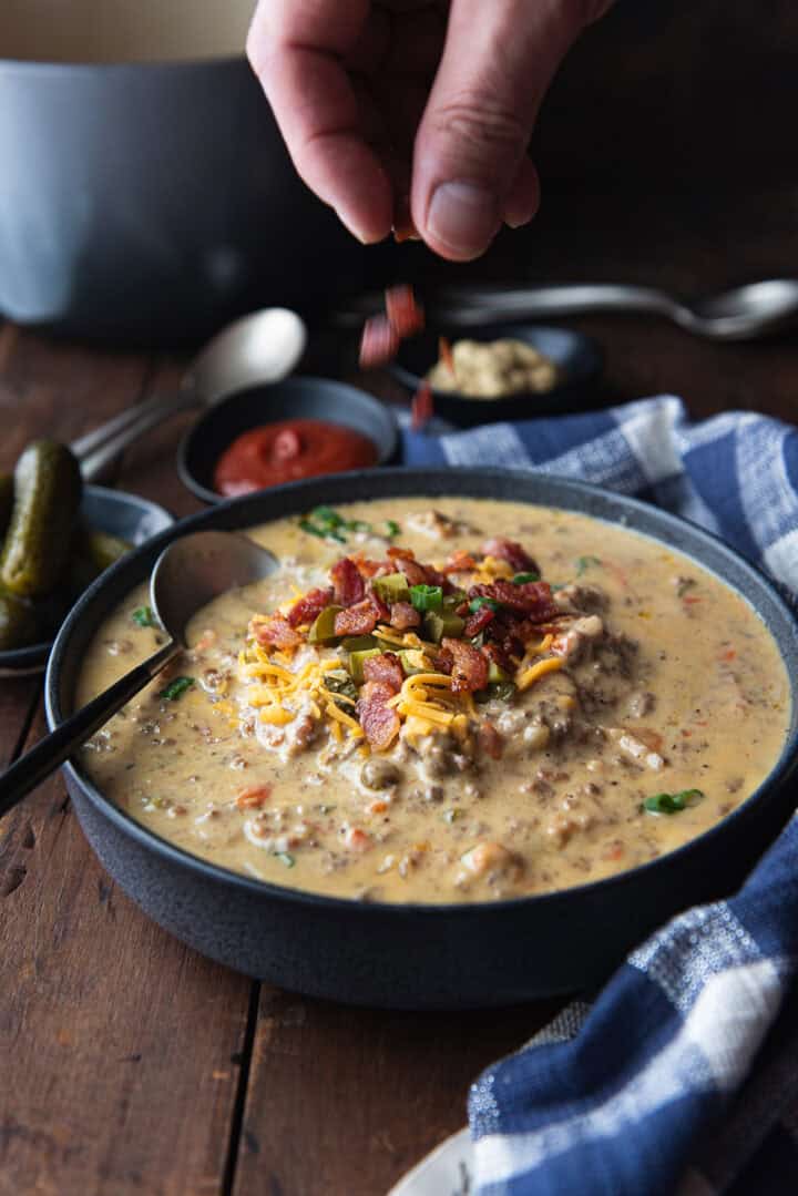 hand sprinkling bacon bits on top of a finished bowl of cheeseburger soup on a rustic wooden table with blue check napkin.