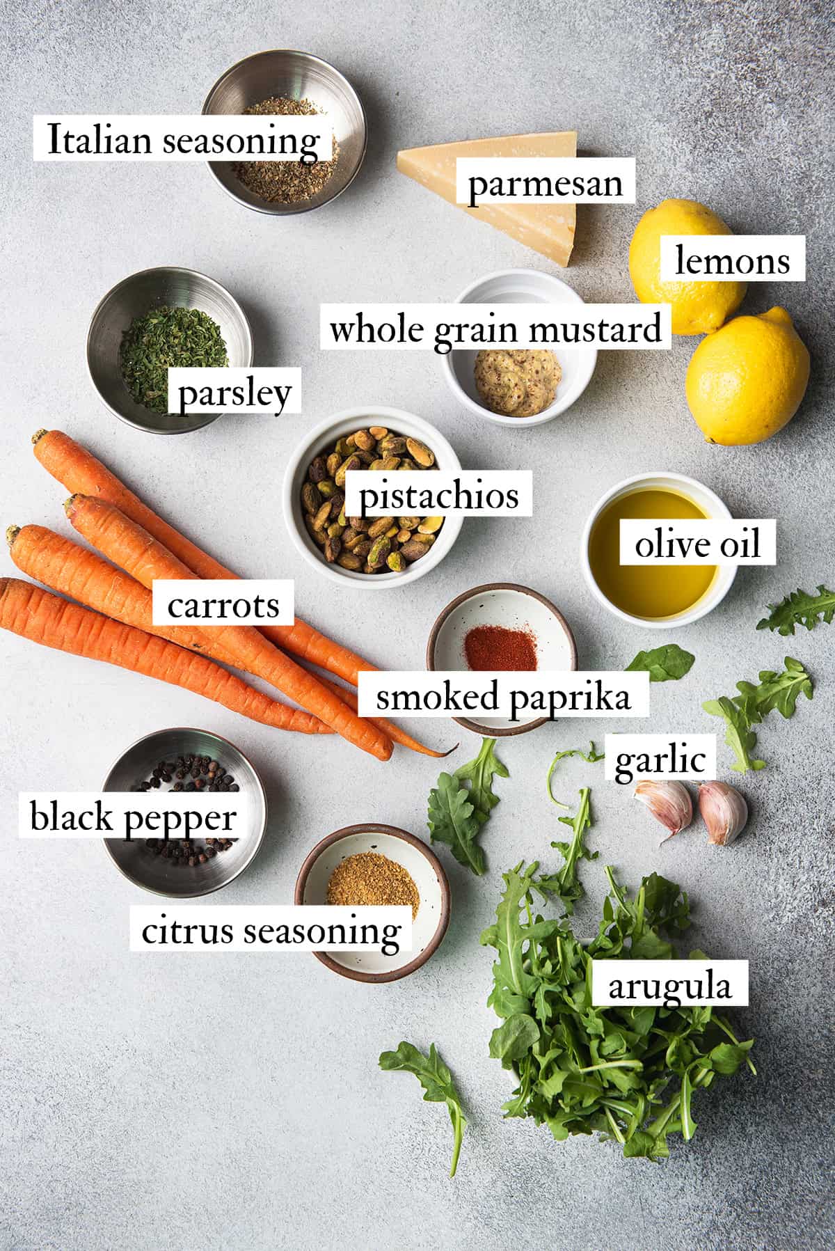 Ingredients for roasted carrot salad including carrots, arugula, pistachios, parmesan, lemon, and spices arranged on a gray surface