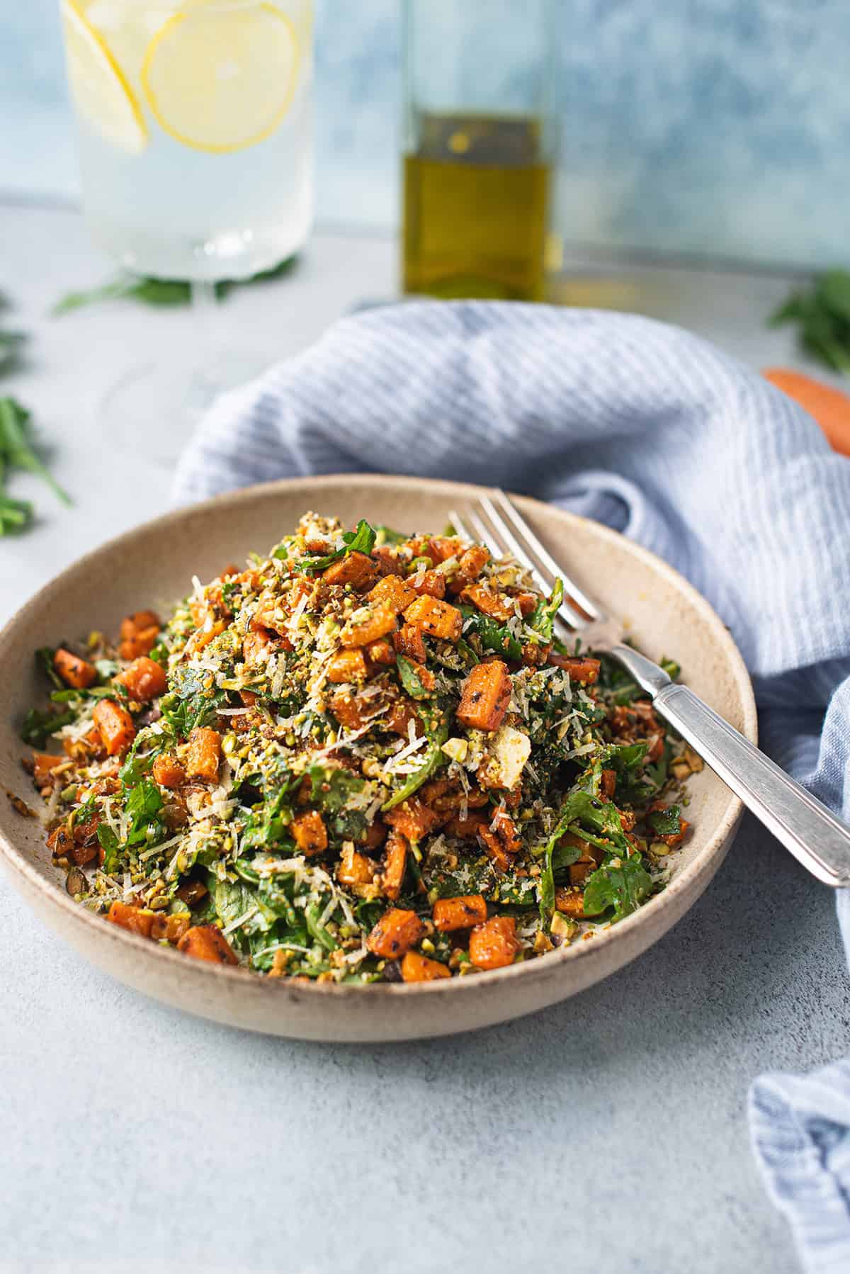 A bowl of roasted carrot salad with arugula and pistachios served with a fork alongside a glass of lemon water