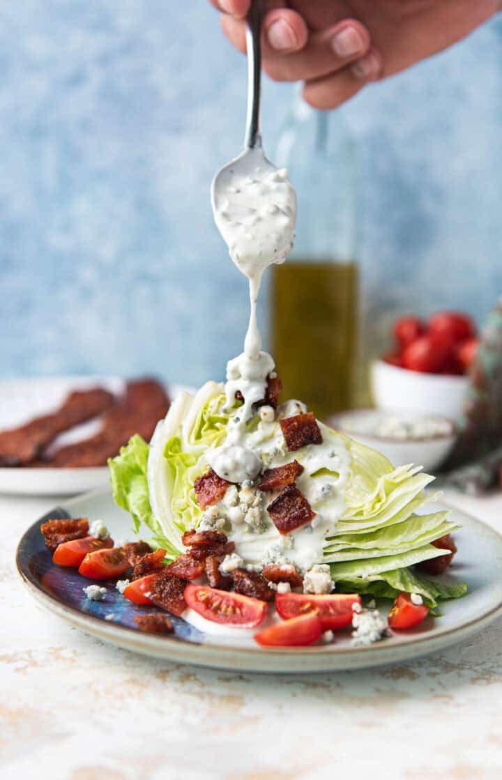 Blue cheese dressing being spooned over a wedge salad topped with candied bacon and tomatoes