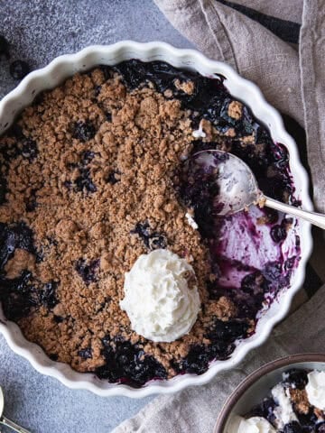 Overhead view of blueberry crumble with a portion scooped out to show the jammy berry filling, topped with whipped cream.